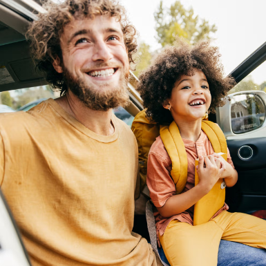 Man and child with backpacks smiling while sitting in a car with open doors.