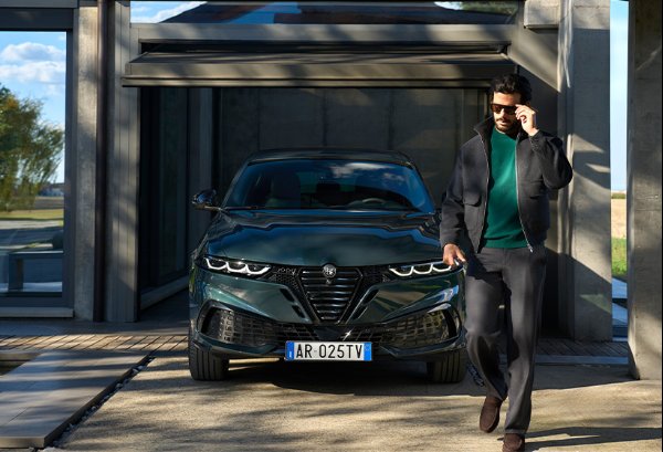 A man in sunglasses walks past a dark green car in a garage with a glass door.