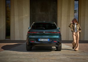 Green car rear view with a woman in a jacket and trousers walking nearby.
