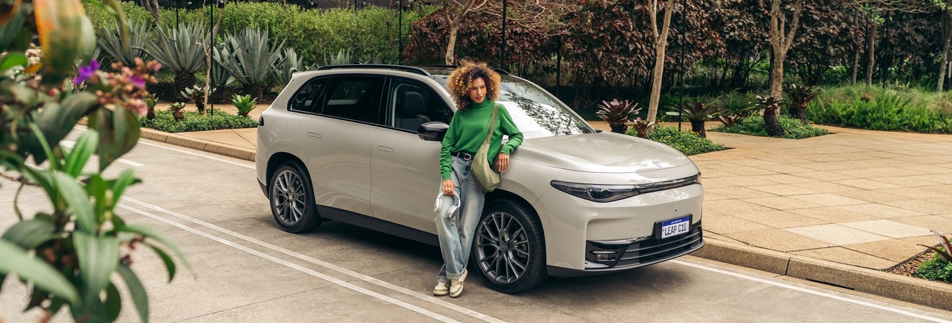 Woman leaning on a white SUV holding a charging cable, surrounded by greenery.