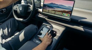 Person in a car touching a smartphone on the console. Large display screen shows landscape image.